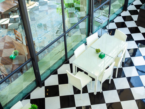 Vista superior de sillas de tejido blanco y mesa de vidrio blanco en el suelo de rejilla blanco y negro en el restaurante. Mesa de comedor moderna en el interior de la cafetería de la casa de cristal
. - Foto, imagen