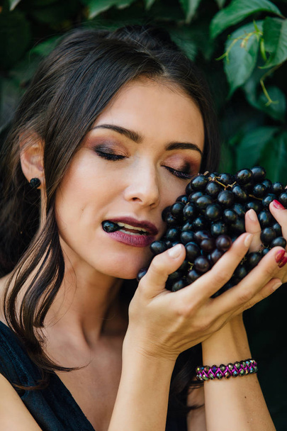 Portrait of young sexy brunette in black dress biting black grapes. - Foto, Imagem