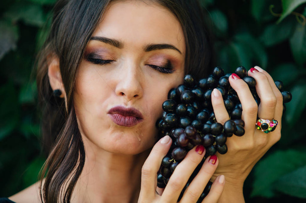Portrait of young sexy brunette in black dress biting black grapes. - Фото, изображение