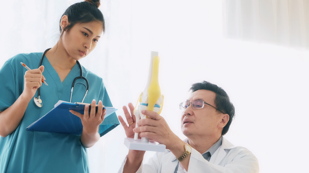 Senior asian man medical doctor sitting on his desk, talking to young medical woman doctor examine bone mock up, taking notes. Young medical doctor career concept. - Footage, Video