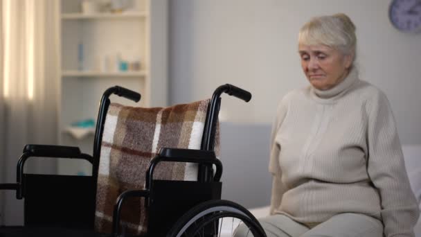 Upset elderly woman sitting on sofa near wheelchair in rehabilitation center - Filmati, video