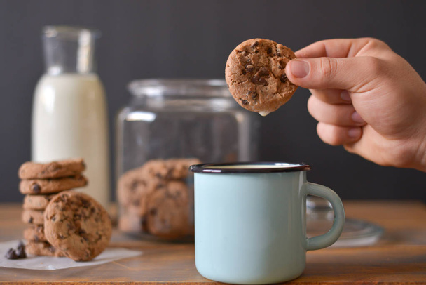 chocolate chip cookies in glass jar with glass bottle of milk and turquoise enamel mug on wooden rustic background with men hand holding one cookie - 写真・画像