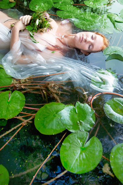 Joven mujer infeliz sexy Ofelia con el pelo rojo rizado se encuentra con las flores con los ojos cerrados en vestido blanco muriendo en el agua en el lago
 - Foto, imagen