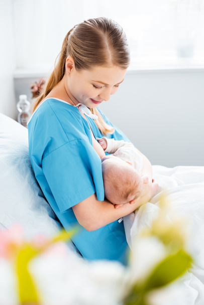 visão lateral de sorrir jovem mãe amamentando bebê recém-nascido na cama no quarto do hospital
  - Foto, Imagem