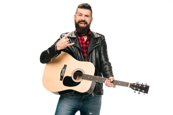 handsome smiling musician in black leather jacket holding acoustic guitar and showing rock and roll sign, isolated on white - Fotó, kép