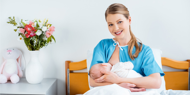feliz joven madre sentada en la cama y sonriendo a la cámara mientras amamanta al bebé recién nacido en la habitación del hospital
 - Foto, imagen