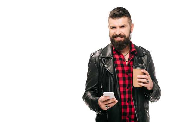 Lächelnder bärtiger Mann in schwarzer Lederjacke mit Coffee to go und Smartphone, isoliert auf weiß - Foto, Bild