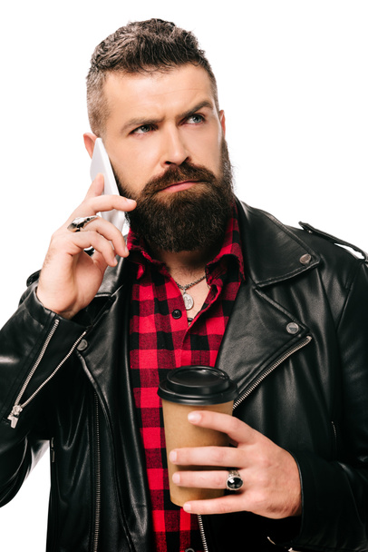 красивий бородатий чоловік в чорній шкіряній куртці, що тримає каву, щоб піти і розмовляти на смартфоні, ізольований на білому
 - Фото, зображення