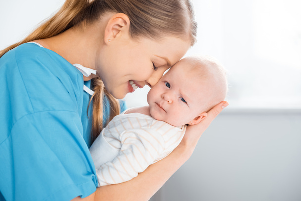 vista lateral de feliz madre joven llevando y abrazando adorable bebé mirando a la cámara en la habitación del hospital
 - Foto, imagen