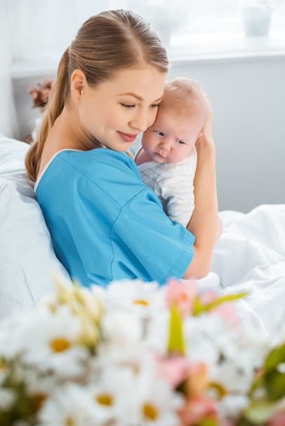 επιλεκτική εστίαση του happy νεαρή μητέρα κάθεται στο κρεβάτι του νοσοκομείου και αγκαλιάζει αξιολάτρευτο μωρό   - Φωτογραφία, εικόνα