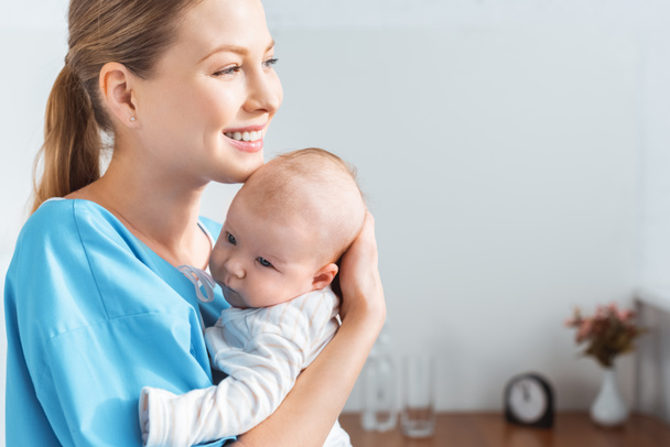 vista lateral da jovem mãe feliz segurando bebê adorável e olhando para longe no quarto do hospital
 - Foto, Imagem