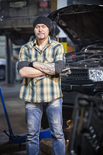 Car repair service. Brutal tired mechanic man stands next to the tool case and holding a wrench - Foto, Imagen