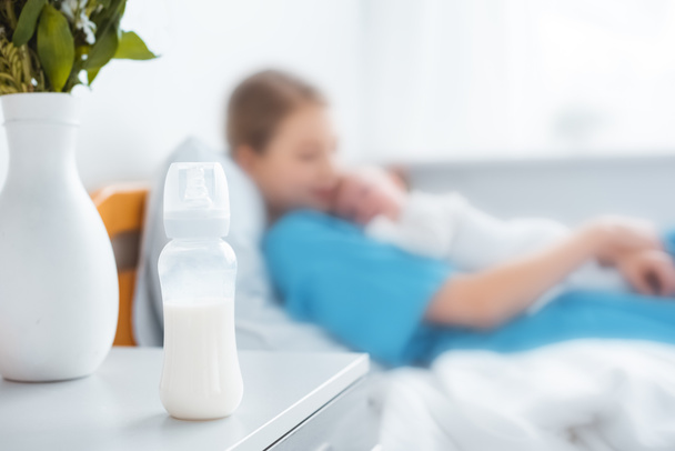 close-up view of baby bottle with milk, vase and mother with newborn baby lying on hospital bed behind - Фото, изображение