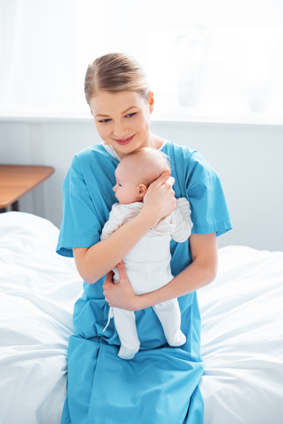 korkea kulma näkymä onnellinen nuori äiti istuu sairaalassa sängyssä ja kuljettaa ihana vastasyntynyt vauva
 - Valokuva, kuva