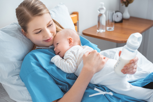 high angle view of smiling young mother holding baby bottle with milk and lying in bed with adorable baby - Фото, изображение