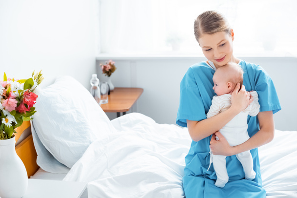 病室で赤ちゃんを保持してベッドの上に座って若い母のハイアングル  - 写真・画像