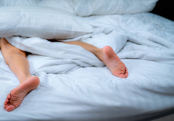 Close-up vrouw blote voeten op het bed over witte deken en bed blad in de slaapkamer thuis of in hotel. Slapen en ontspannen concept. Luie ochtend. Blote voeten van vrouw liggend op witte comfort bed en dekbed. - Foto, afbeelding