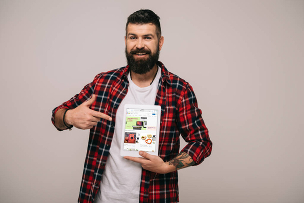 γενειοφόρος χαμογελαστός άνθρωπος σε καρώ πουκάμισο δείχνει στο ψηφιακό tablet με το app ebay, απομονώνονται σε γκρι - Φωτογραφία, εικόνα