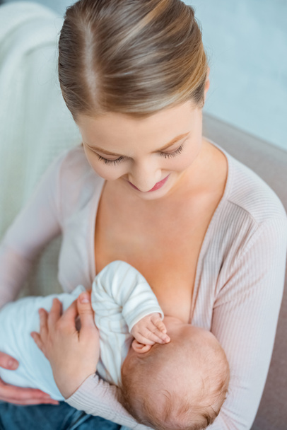 high angle view of beautiful smiling young woman sitting on couch and breastfeeding infant daughter - Photo, image