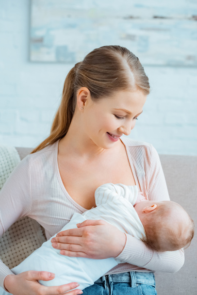 beautiful smiling young woman sitting on couch and breastfeeding infant daughter - Photo, Image