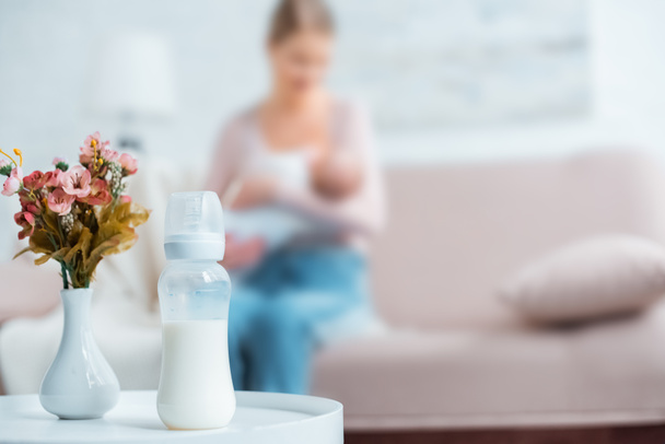 close-up view of baby bottle with milk, flowers in vase and mother breastfeeding baby behind at home - Foto, Imagem