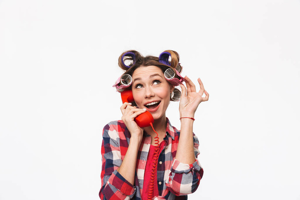 Cheerful housewife with curlers in hair standing isolated over white background, talking on a landline phone - Foto, Bild