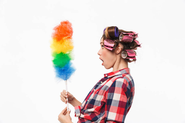 Busy housewife with curlers in hair standing isolated over white background, holding duster - Foto, afbeelding