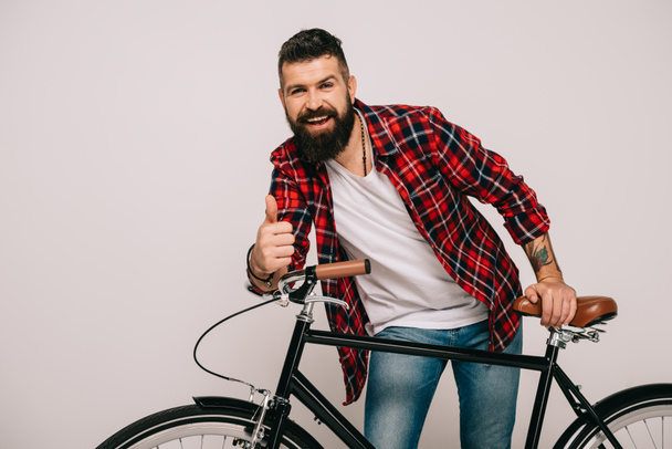  glimlachend bebaarde man poseren met de fiets en het tonen van de duim omhoog geïsoleerd op grijs - Foto, afbeelding