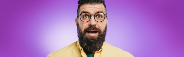 shocked bearded man in eyeglasses looking at camera, isolated on purple - 写真・画像