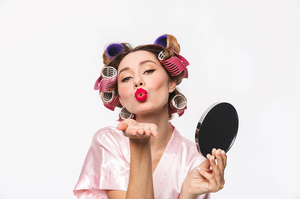 Beautiful housewife with curlers in hair wearing robe standing isolated over white background, looking at herself at the mirror, sending kiss - Фото, зображення