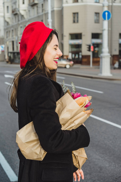 Young beautiful girl Frenchwoman brunette in a red beret and a black coat goes along the street of the European city with a bouquet of flowers and French baguettes. A happy smile and a charming look, the French style of Paris - Foto, afbeelding