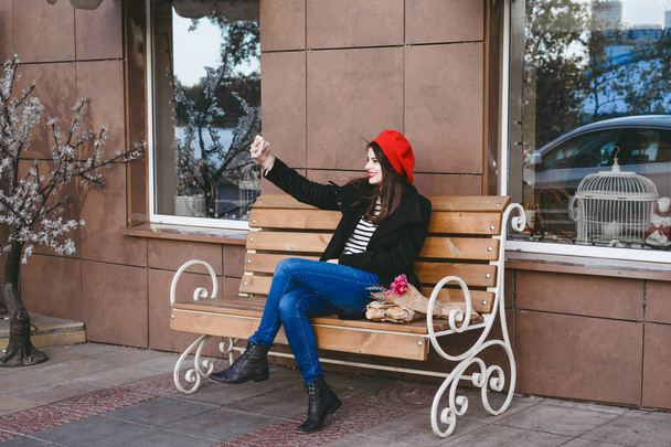 beautiful brunette woman in red beret sits on the street on the bench, makes selfie, uses a smartphone - Valokuva, kuva