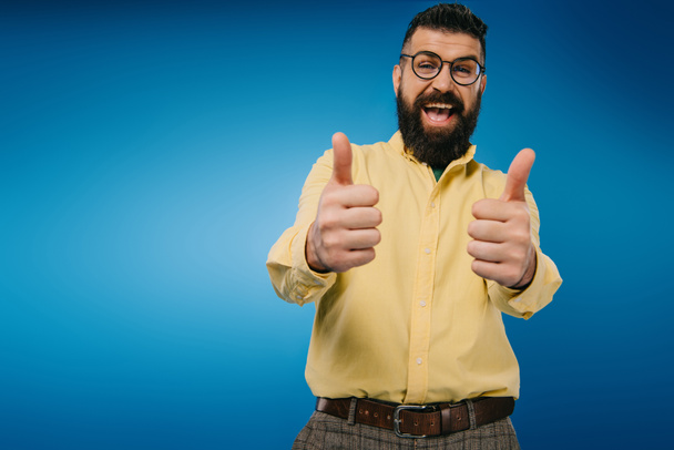 excited bearded man in eyeglasses showing thumbs up isolated on blue - Photo, Image