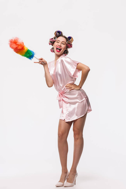 Full length portrait of a pretty housewife with curlers in her hair wearing robe standing isolated over white background, posing with a duster - Photo, Image