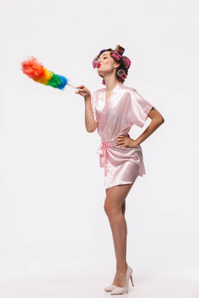 Full length portrait of a pretty housewife with curlers in her hair wearing robe standing isolated over white background, posing with a duster - Foto, Imagem
