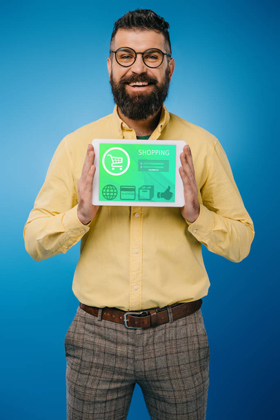 smiling bearded man showing digital tablet with shopping app, isolated on blue - Photo, Image
