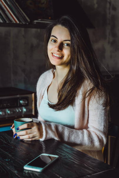 μια όμορφη μελαχρινή κοπέλα με ένα όμορφο χαμόγελο και εκφραστικά μάτια καστανά, πίνοντας καφέ και χρησιμοποιώντας ένα smartphone - Φωτογραφία, εικόνα