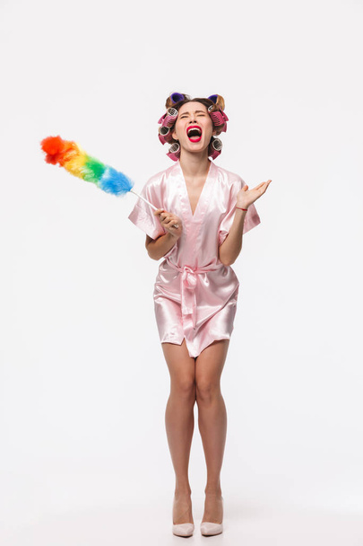 Full length portrait of a pretty angry housewife with curlers in her hair wearing robe standing isolated over white background, posing with a duster - Foto, afbeelding