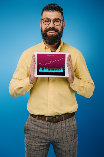 smiling bearded man presenting digital tablet with tariding app, isolated on blue - Fotoğraf, Görsel