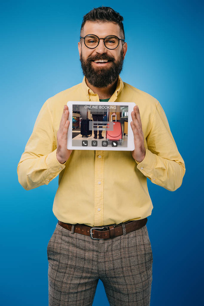 улыбающийся бородатый мужчина представляет цифровой планшет с приложением онлайн бронирования, изолированный на синий
 - Фото, изображение