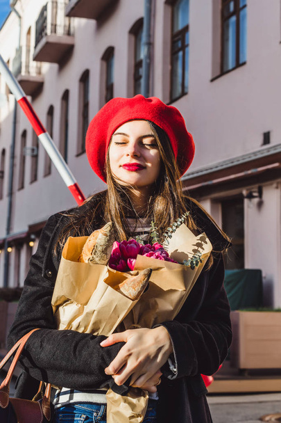Young beautiful girl Frenchwoman brunette in a red beret and a black coat goes along the street of the European city with a bouquet of flowers and French baguettes. A happy smile and a charming look, the French style of Paris - Φωτογραφία, εικόνα