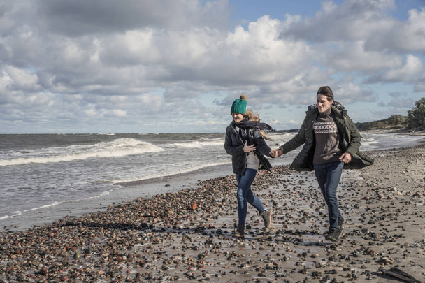a young couple in love spending weekends on the coast of the cold Baltic sea, sandy beach, warm jackets, family weekend man and woman running on the beach. - Фото, изображение