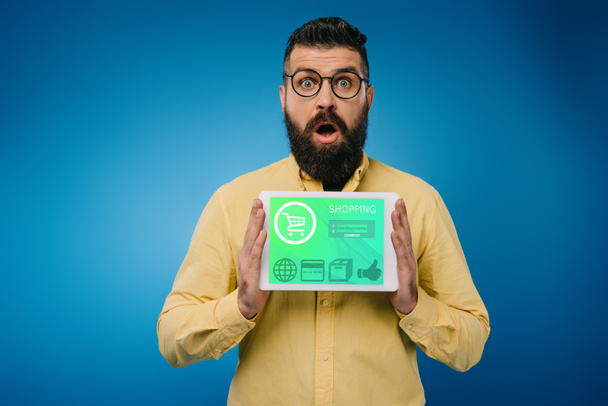 homem barbudo chocado apresentando tablet digital com aplicativo de compras, isolado em azul
 - Foto, Imagem
