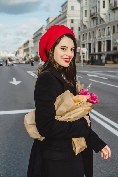 Young beautiful girl Frenchwoman brunette in a red beret and a black coat goes along the street of the European city with a bouquet of flowers and French baguettes. A happy smile and a charming look, the French style of Paris - Foto, Imagen