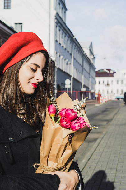 Young beautiful girl Frenchwoman brunette in a red beret and a black coat goes along the street of the European city with a bouquet of flowers and French baguettes. A happy smile and a charming look, the French style of Paris - 写真・画像