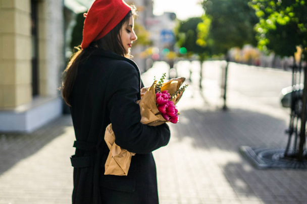 Young beautiful girl Frenchwoman brunette in a red beret and a black coat goes along the street of the European city with a bouquet of flowers and French baguettes. A happy smile and a charming look, the French style of Paris - Valokuva, kuva