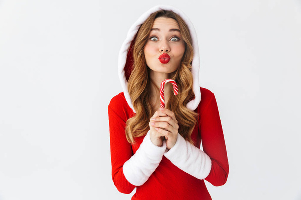 Cheerful young girl wearing Christmas dress standing isolated over white background, eating Christmas candy treats - Fotoğraf, Görsel