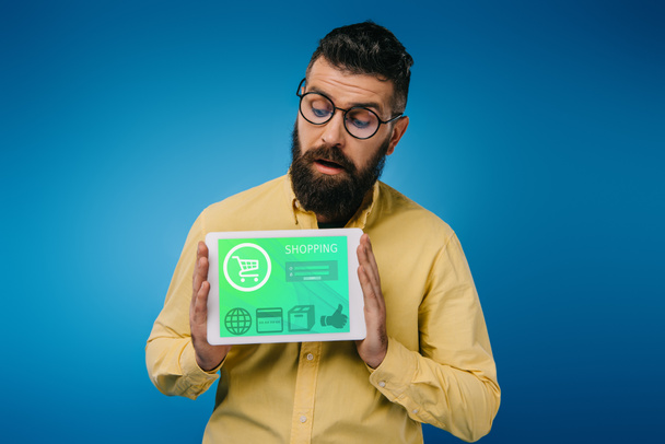 зацікавлений бородатий чоловік дивиться на цифровий планшет з додатком для покупок, ізольований на синьому
 - Фото, зображення