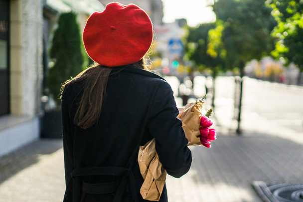 Young beautiful girl Frenchwoman brunette in a red beret and a black coat goes along the street of the European city with a bouquet of flowers and French baguettes. A happy smile and a charming look, the French style of Paris - Φωτογραφία, εικόνα