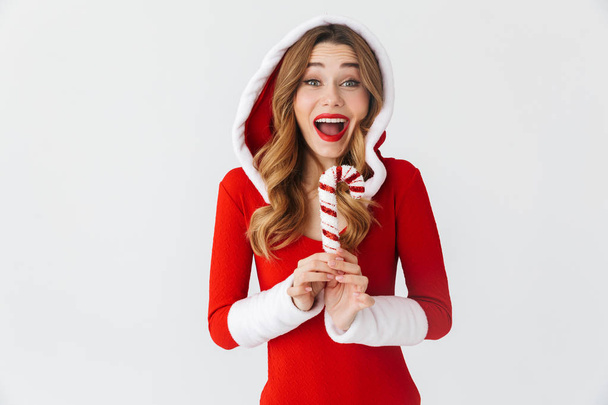 Cheerful young girl wearing Christmas dress standing isolated over white background, eating Christmas candy treats - Fotoğraf, Görsel